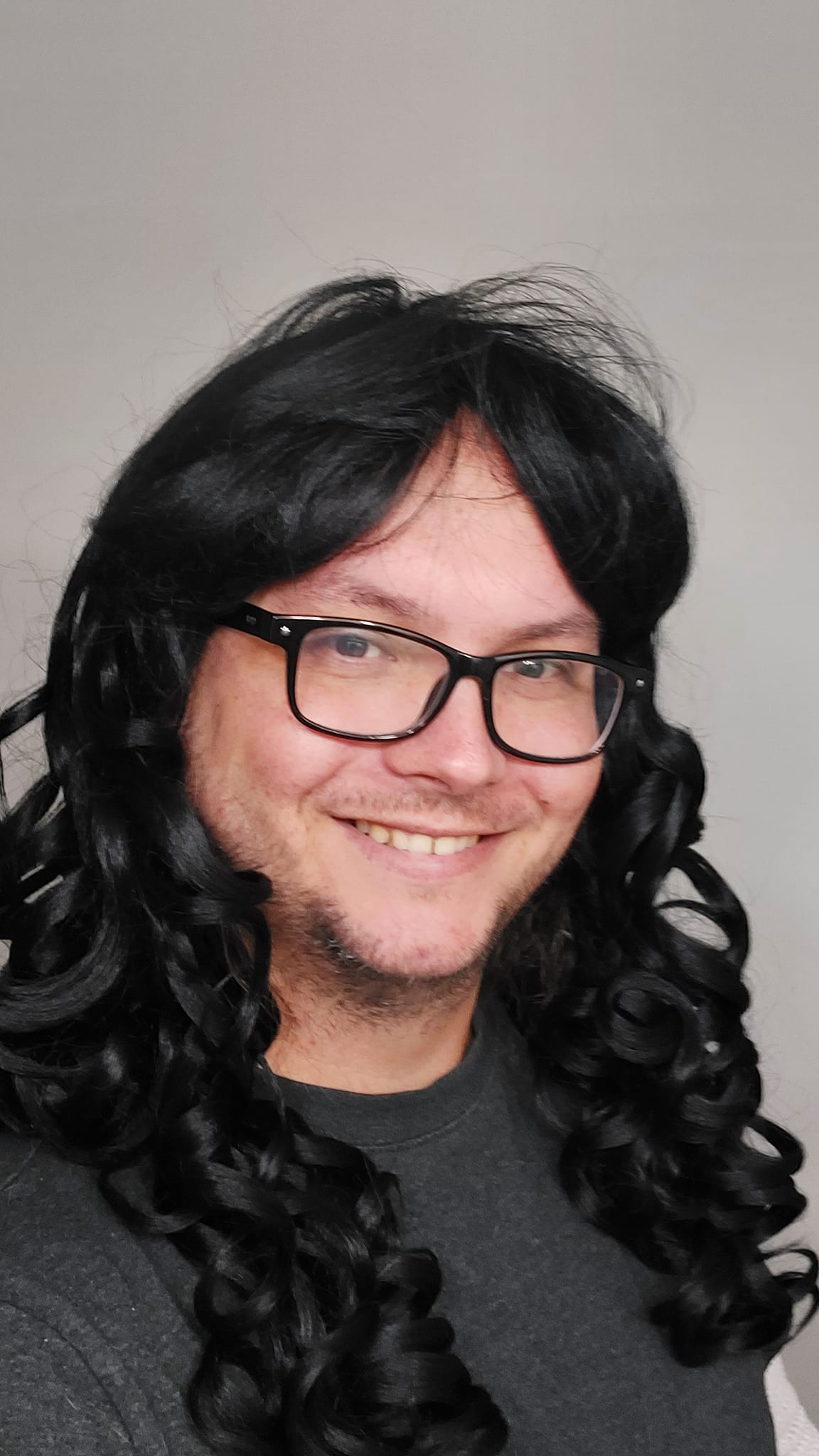 A man wearing a black shirt wears a long black curly costume wig